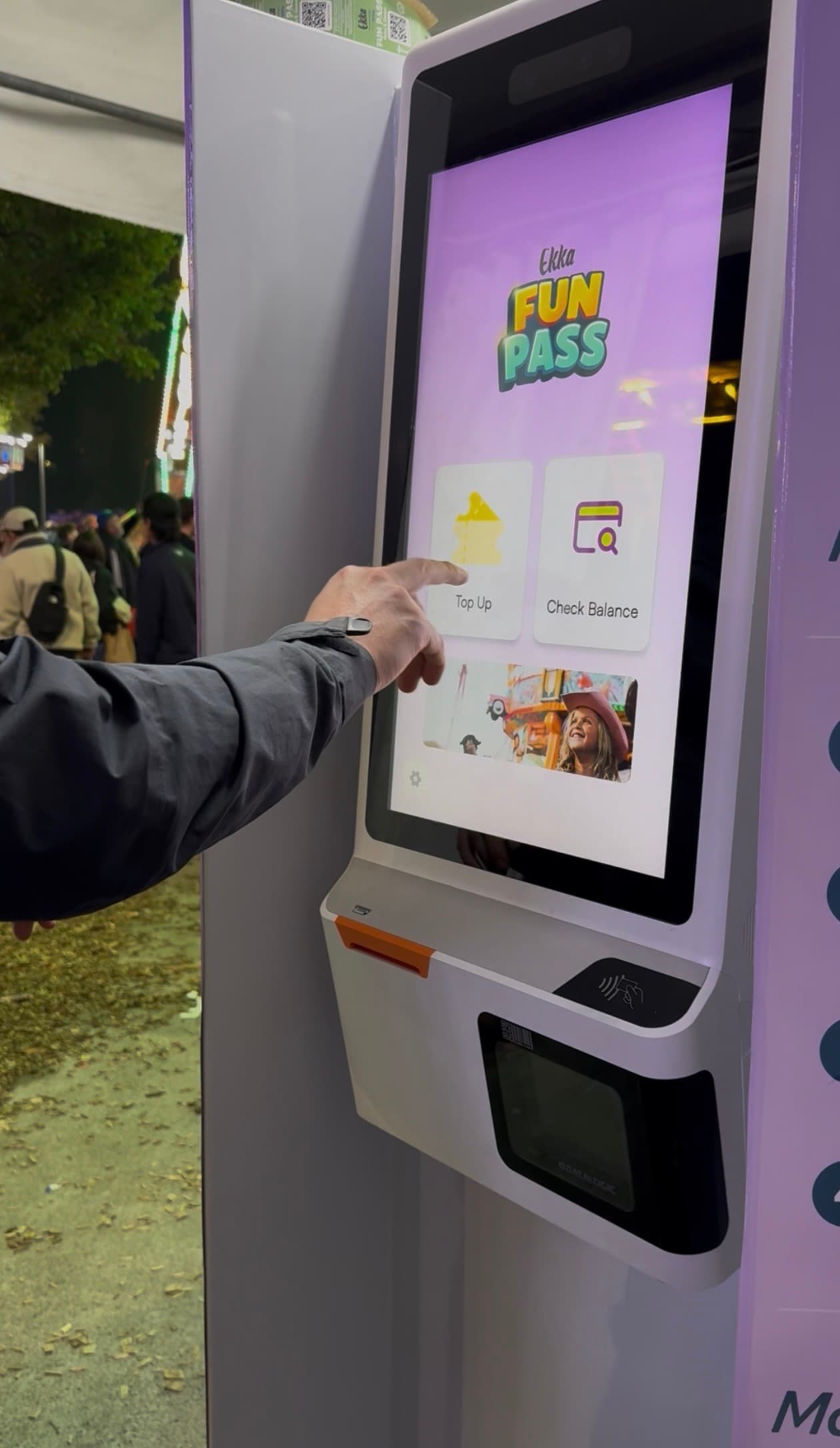Close-up of a visitor tapping the Ekka Fun Pass kiosk touchscreen at night